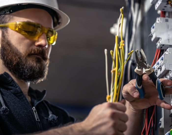 A male electrician works in a switchboard with an electrical connecting cable, connects the equipment with tools.